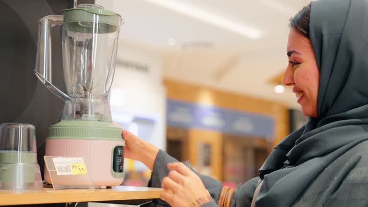 مقارنة المنتجات وقراءة التفاصيل قبل الشراء، تصوير مقرب لامرأة عربية خليجية إماراتية محجبة داخل متجر للأجهزة الكهربائية تمسك جهاز خلاط كهربائي وتتفقده ، ايماءات السعادة و السرور، اختيار وشراء الأجهزة المنزلية