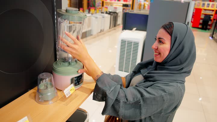 تصوير مقرب لامرأة عربية خليجية إماراتية محجبة داخل متجر للأجهزة الكهربائية تمسك جهاز خلاط كهربائي وتتفقده ، ايماءات السعادة و السرور، مقارنة المنتجات وقراءة التفاصيل قبل الشراء، اختيار وشراء الأجهزة المنزلية