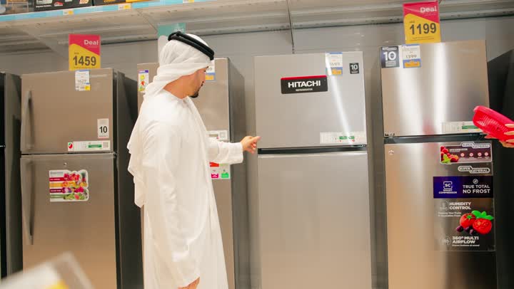 Discussion and selection of a product before purchase, an Emirati Gulf Arab woman in a hijab stands in front of display refrigerators inside an electrical appliances store with her husband, holding a red fan. A family shopping atmosphere, choosing and buying household appliances, gestures of happiness and joy.