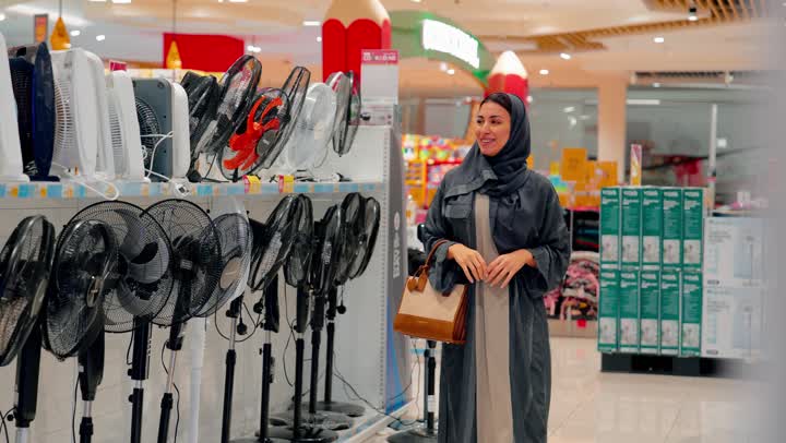 تقييم المنتجات ومقارنتها، التسوق داخل متجر إلكترونيات، اختيار أجهزة التهوية المناسبة، امراة عربية خليجية اماراتية ترتدي العباءة والحجاب  تقوم بتفقد مراوح كهربائية منزلية معروضة داخل المتجر، مفهوم الشراء الواعي، نمط الحياة العصرية