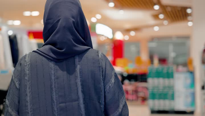 اختيار أجهزة التهوية المناسبة، تصوير من الخلف لامرأة عربية خليجية اماراتية ترتدي العباءة والحجاب  تقوم بتفقد مراوح كهربائية منزلية معروضة داخل المتجر، مقارنة المراوح قبل الشراء، التسوق داخل متجر إلكترونيات، مفهوم الشراء الواعي، نمط الحياة العصرية