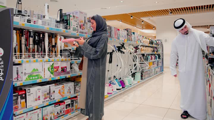 مقارنة أدوات المطبخ قبل الشراء، امرأة عربية خليجية اماراتية ترتدي العباءة والحجاب تقوم بفحص خلاط يدوي كهربائي برفقة زوجها، متجر أجهزة منزلية حديث، اختيار الأجهزة المناسبة، التسوق داخل متجر إلكترونيات، مفهوم الشراء الواعي