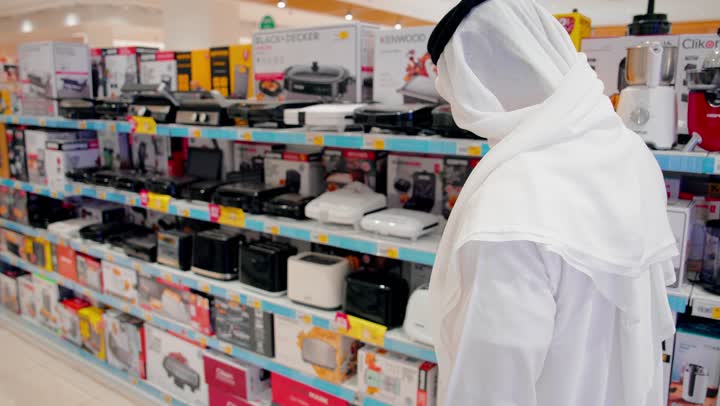 تجربة الأجهزة المعروضة، مفهوم التسوق العائلي، امرأة عربية خليجية اماراتية ترتدي العباءة والحجاب تقوم بفحص توستر كهربائي برفقة زوجها، مقارنة المنتجات قبل الشراء، التسوق داخل متجر إلكترونيات