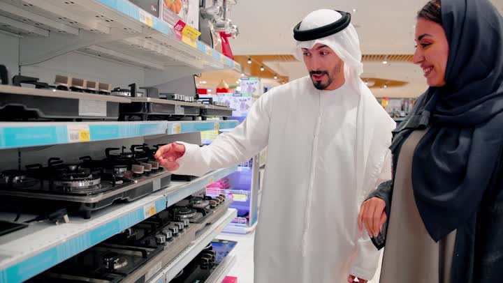 مفهوم التسوق العائلي،  اختيار الأجهزة المنزلية المناسبة، رجل عربي خليجي يرتدي الزي التقليدي يشير إلى موقد غاز معروض داخل متجر أجهزة منزلية حديث برفة زوجته،ايماءات السعادة و السرور