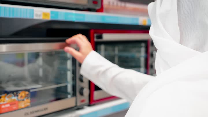 مقارنة الأجهزة المنزلية،  فحص التفاصيل قبل اتخاذ قرار الشراء، التسوق داخل متجر إلكترونيات، تصوير مقرب لرجل عربي خليجي يرتدي الزي التقليدي يقوم بتفقد فرن كهربائي، اختيار الفرن المناسب، مفهوم الشراء الواعي