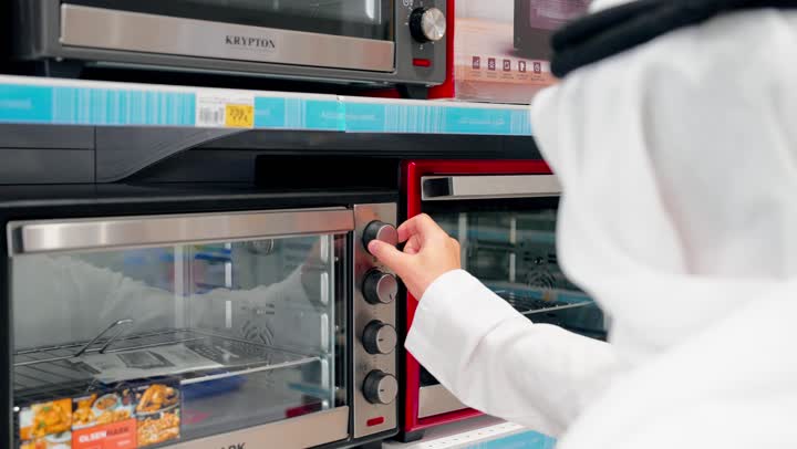 فحص التفاصيل قبل اتخاذ قرار الشراء، التسوق داخل متجر إلكترونيات، تصوير مقرب لرجل عربي خليجي يرتدي الزي التقليدي يقوم بتفقد فرن كهربائي، مقارنة الأجهزة المنزلية، اختيار الفرن المناسب، مفهوم الشراء الواعي