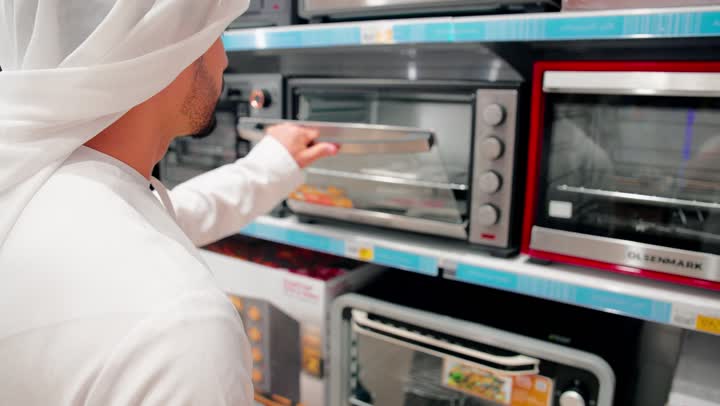 Shopping inside an electronics store, a close-up shot of a Gulf Arab man wearing traditional attire inspecting an electric oven, checking the details before making a purchase decision, comparing home appliances, choosing the right oven, the concept of conscious buying.