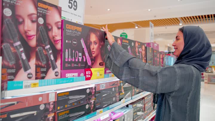 مفهوم التسوق النسائي، امرأة عربية خليجية  اماراتية ترتدي العباءة والحجاب تقوم باختيار وشراء جهاز تصفيف شعر، تفحص المنتجات المعروضة قبل اتخاذ قرار الشراء، اقتناء أجهزة العناية الشخصية، تنوع منتجات التجميل والعناية، التسوق داخل المتاجر الحديثة