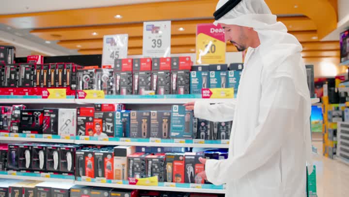 اتخاذ قرار الشراء المناسب، مقارنة المنتجات الإلكترونية، رجل عربي خليجي اماراتي يرتدي الزي التقليدي يقف داخل متجر إلكترونيات ، اختيار وشراء منتجات عناية شخصية ، مفهوم التسوق داخل المتاجر، تنوع ملحقات العناية الشخصية