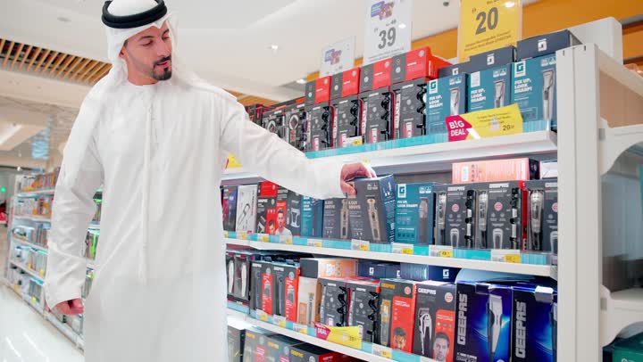 مقارنة المنتجات الإلكترونية، رجل عربي خليجي اماراتي يرتدي الزي التقليدي يقف داخل متجر إلكترونيات ، اختيار وشراء منتجات عناية شخصية ، مفهوم التسوق داخل المتاجر، تنوع ملحقات العناية الشخصية، اتخاذ قرار الشراء المناسب