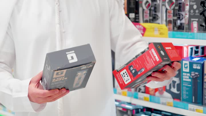 تنوع الإكسسوارات الإلكترونية، مقارنة المواصفات التقنية للمنتجات، تصوير مقرب لرجل عربي خليجي امارتي يرتدي الزي التقليدي يمسك عبوتين لمنتجات إلكترونية داخل متجر إلكترونيات ، مفهوم التسوق الذكي، اقتناء منتجات تقنية حديثة