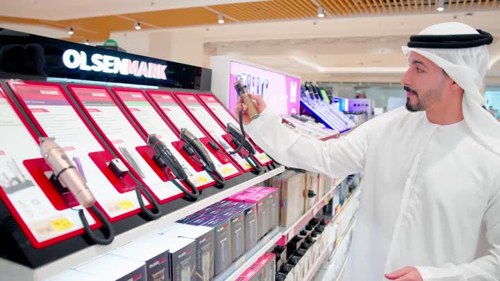 اختيار الأجهزة المناسبة للاستخدام اليومي، رجل عربي خليجي اماراتي يرتدي الزي التقليدي يقف داخل متجر إلكترونيات ، أثناء اختيار وشراء ماكينة حلاقة كهربائية، مفهوم التسوق الشخصي، مقارنة ملحقات العناية الشخصية، تنوع المنتجات الإلكترونية