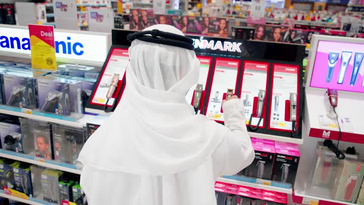 مقارنة المنتجات التقنية الصغيرة، تصوير مقرب من الخلف لرجل عربي خليجي يرتدي الزي التقليدي يقف داخل متجر إلكترونيات أثناء اختيار وشراء ملحقات إلكترونية، اختيار الإكسسوارات الإلكترونية المناسبة، تنوع المنتجات الرقمية