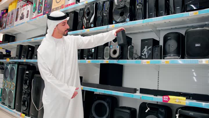 اختيار ملحقات إلكترونية حديثة، اقتناء أجهزة إلكترونية منزلية، رجل عربي خليجي يرتدي الزي التقليدي يمسك مكبر صوت إلكتروني ، اختيار وشراء جهاز صوتي مناسب، مفهوم التسوق داخل المتاجر، مقارنة الأجهزة الصوتية، جودة الصوت والتقنيات الحديثة