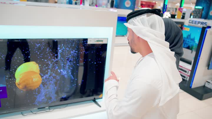 زوجان عربيان خليجيان يرتديان الزي التقليدي يقفان أمام شاشة تلفاز ذكية داخل متجر إلكترونيات ويقومان بمشاهدة محتوى معروض، مفهوم تجربة الشاشات الذكية، مقارنة جودة العرض والصورة، اختيار الأجهزة المنزلية الحديثة، تطور تقنيات العرض الرقمية