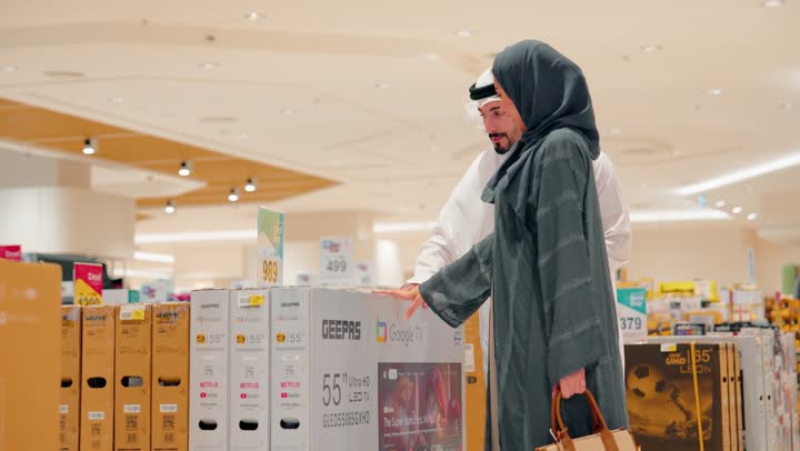 The concept of choosing modern home appliances, an Arab Gulf Emirati woman wearing an abaya and hijab stands inside an electronics store with her husband, pointing to displayed smart TVs, comparing the sizes of the smart screens.