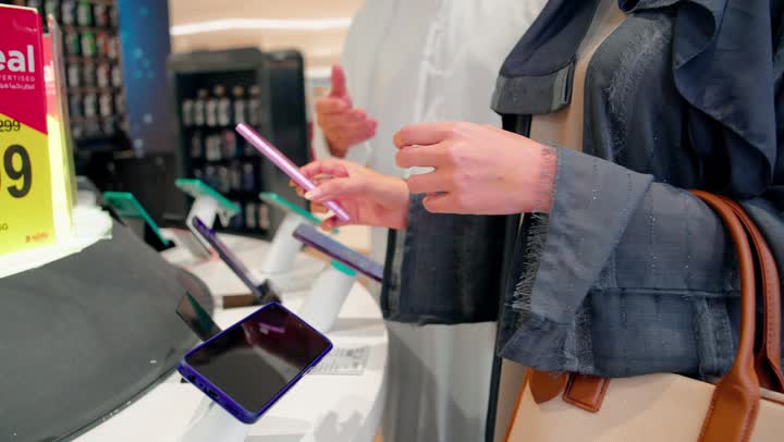 تجربة استخدام الهواتف الذكية، مفهوم التسوق المشترك، تصوير مقرب لامراه عربيه خليجيه اماراتية ترتدي العباءة و الحجاب تقوم بتجربة هاتف ذكي داخل متجر إلكترونيات حديث برفقة زوجها، تطور التكنولوجيا الرقمية