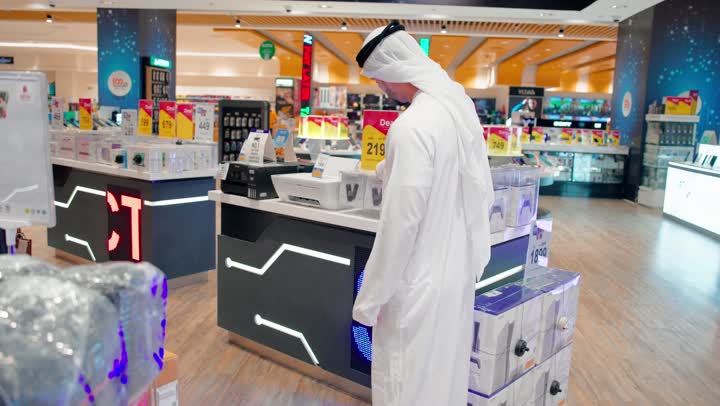 مقارنة أسعار الهواتف الذكية، رجل عربي خليجي اماراتي يرتدي الكندورة و الغتره يسيرداخل متجر إلكترونيات حديث برفقة زوجته، مفهوم التسوق داخل المتاجر، اختيار الأجهزة التقنية المناسبة، تطور التكنولوجيا الرقمية