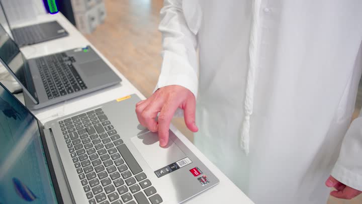 Comparison of technical specifications, a close-up shot of the hand of an Arab Gulf man wearing traditional attire testing laptop devices displayed inside a modern electronics store, the concept of tech shopping, choosing portable computers, using modern technology.