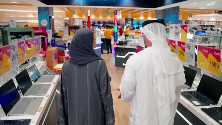عروض وخصومات المنتجات الإلكترونية، تصوير من الخلف لزوجان عربيان خليجيان يرتديان الزي التقليدي يسيران داخل متجر إلكترونيات يقومان بتفقد الأجهزة المعروضة، مفهوم التسوق العائلي، اختيار الأجهزة التقنية الحديثة، تطور التكنولوجيا الرقمية
