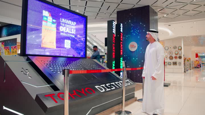 Exploring the latest devices and digital technologies, a rear view of an Arab Gulf Emirati woman wearing an abaya and hijab standing in front of a large display screen and a laptop inside a tech store with her husband, the evolution of modern technologies and the selection of smart devices.