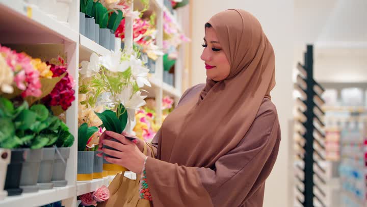 امرأة عربية خليجية إماراتية محجبة ترتدي العباءة داخل متجر ديكور منزلي تقوم بتأمل واختيار نبتة زهور صناعية بعناية، اختيار قطع الزينة، ايماءات السعادة والرضا أثناء التسوق، مفهوم الاهتمام بتنسيق المنزل