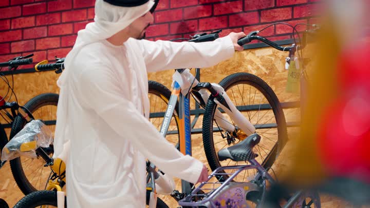رجل عربي خليجي اماراتي  يرتدي الكندورة و الغترة يقوم بتفقد دراجات هوائية معروضة داخل المتجر، أنشطة ترفيهية شبابية، ممارسة الهوايات الرياضية الحديثة، الاهتمام بنمط الحياة الصحي، اختيار المعدات الرياضية المناسبة
