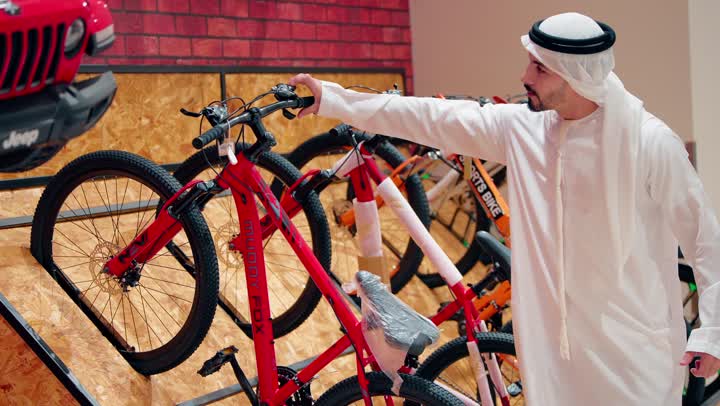 Youth recreational activities, an Emirati Gulf Arab man wearing a kandura and ghutrah is inspecting bicycles displayed inside the store, practicing modern sports hobbies, focusing on a healthy lifestyle, and choosing the appropriate sports equipment.