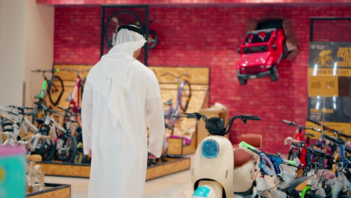 Interest in sports hobbies, a rear view of an Arab Gulf Emirati man wearing a kandura and ghutrah standing inside a bicycle shop, carefully inspecting and selecting bicycles while shopping.