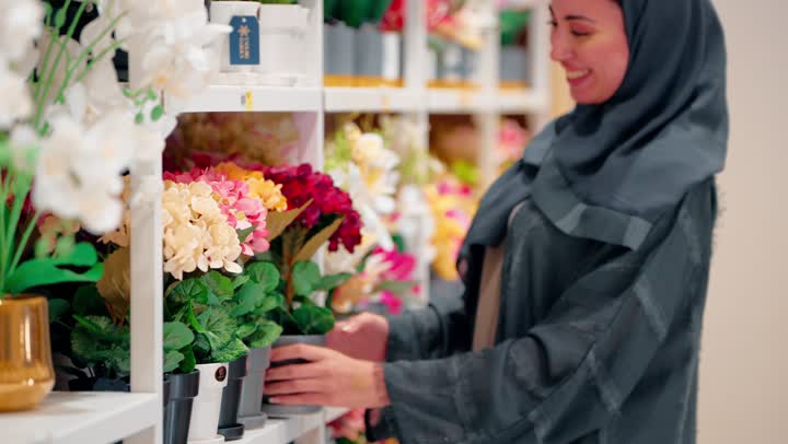 Choosing decorative pieces, an Emirati Gulf Arab woman in a hijab wearing an abaya inside a home decor store carefully contemplating and selecting an artificial flower plant, gestures of happiness and satisfaction while shopping, the concept of caring for home arrangement.