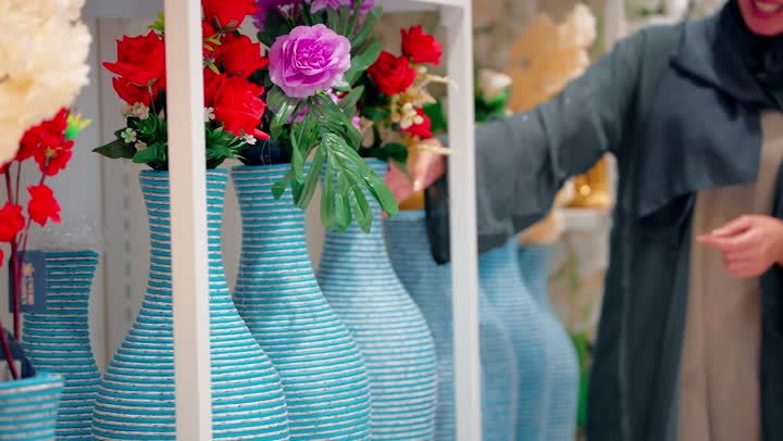 تبادل الآراء أثناء التسوق، امراة عربيه خليجيه اماراتية ترتدي العباءة و الحجاب تقوم بعرض مزهرية ورد صناعي على صديقتها،  تبادل الحديث والإشارات الإيجابية، الاهتمام باختيار القطع الجمالية لتزيين المنزل، مفهوم التسوق النسائي، اختيار الديكور المنزلي