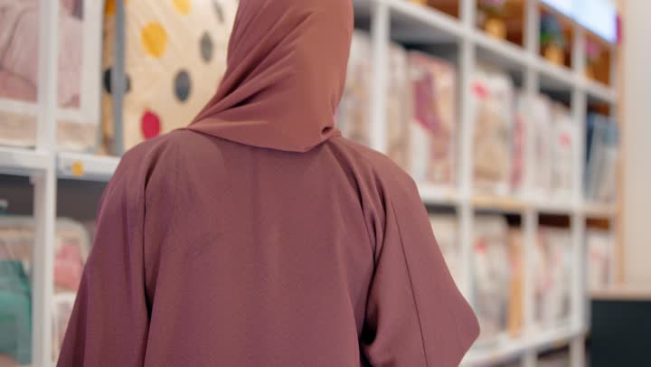 فحص الخامات بعناية والانتباه للتفاصيل، امرأة عربية خليجية إماراتية محجبة ترتدي العباءة تقوم باختيار مفروشات ومنسوجات منزلية، التسوق داخل مركز تجاري حديث، مفهوم تجهيز المنزل، اختيار المستلزمات المنزلية