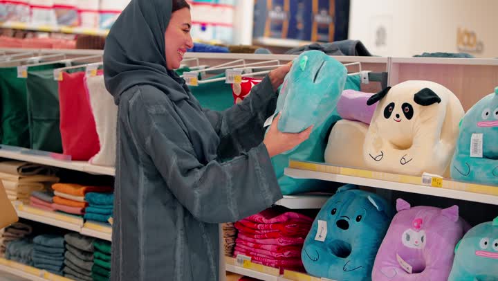 امراه عربيه خليجيه اماراتية محجبة ترتدي العباءة تقوم بعرض وسادة أطفال على صديقتها أثناء التسوق، مفهوم التسوق الأسري، اختيار مستلزمات الأطفال، ايماءات السعادة و السرور