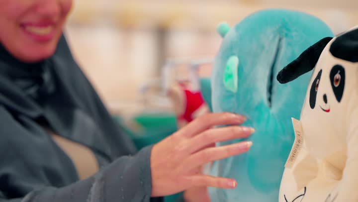 مفهوم تجهيز مستلزمات الطفل، تصوير مقرب لامرأة عربية خليجية اماراتية محجبة تتسوق داخل متجر مستلزمات منزلية تمسك وسادة أطفال تقوم بفحص الخامة والملمس، اختيار منتجات ناعمة وآمنة