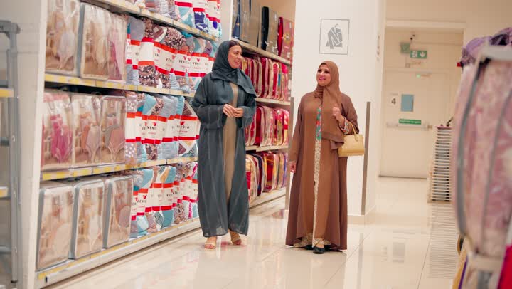 تبادل الحديث أثناء اختيار مفارش وأغطية الأسرة، التعاون الأسري في اختيار المفروشات، امرأتان عربيتان خليجيتان اماراتيان محجبتان ترتديان العباءة تتسوقان داخل متجر مستلزمات منزلية، فحص جودة المنتجات والخامات، مفهوم التسوق العائلي
