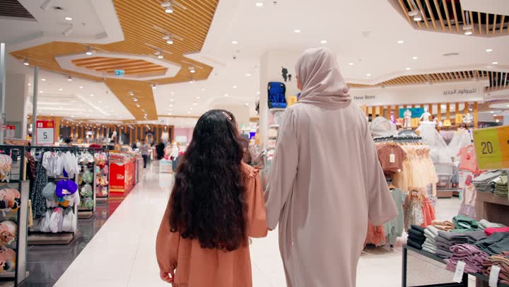 تصوير من الخلف لأم عربية خليجية اماراتية محجبة تمسك يد ابنتها أثناء التسوق داخل متجر ملابس،  نزهة تسوق عائلية داخل مركز تجاري حديث، التجول بين أقسام الملابس