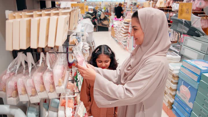اختيار مستلزمات المولود الجديد، مفهوم الاستعداد للأمومة، امرأة عربية خليجيه اماراتية محجبة ترتدي العباءة تقف داخل متجر مستلزمات الأطفال برفقة ابنتها ، ترابط عائلي ومشاعر حب واهتمام