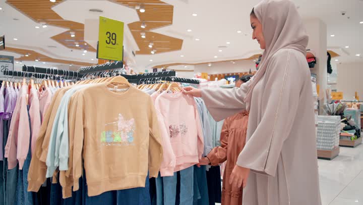 التسوق داخل المولات التجارية، امرأة عربية خليجيه اماراتية محجبة ترتدي العباءة تقوم باختيار ملابس أطفال داخل محل اطفال برفقة ابنتها، الاهتمام بالأطفال واختيار الملابس المناسبة، أجواء عائلية دافئة، مفهوم الأمومة و الترابط الأسري