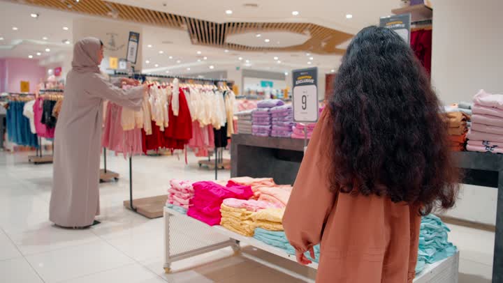 مشاركة الأطفال في اختيار الملابس، بنت عربيه خليجيه اماراتية ترتدي الزي الكاجوال تقوم بعرض فستان على والدتها داخل متجر ملابس في مركز تجاري حديث، متعة التسوق العائلي، الاهتمام بمظهر الأطفال، مفهوم الترابط الأسري