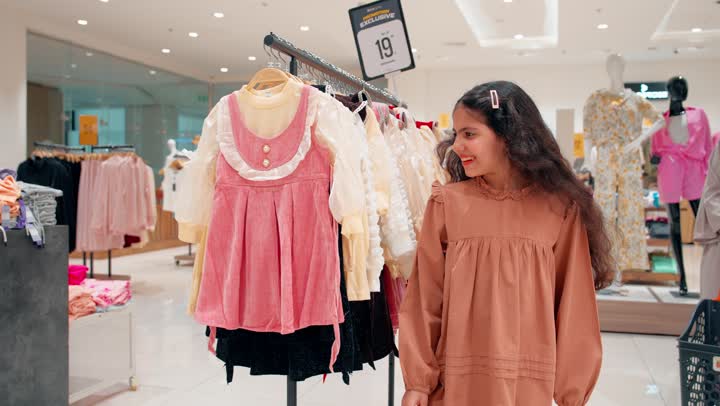مفهوم التسوق العائلي وملابس الأطفال، بنت عربية خليجيه امارتية تقوم باختيار فستان داخل متجر ملابس ، فحص ملابس البنات المعروضة، اهتمام الأطفال بتنسيق الملابس، أجواء تسوق عائلية داخل مركز تجاري حديث