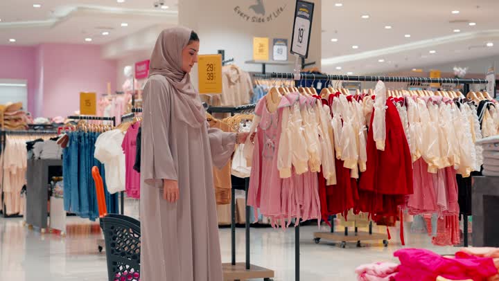العلاقة الأسرية الدافئة بين الأم والطفلة، أم عربية خليجية امارتية محجبة تساعد ابنتها في تجربة الملابس داخل متجر ملابس، مفهوم التسوق العائلي داخل المولات التجارية
