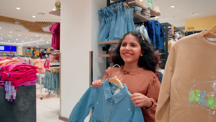 السعادة والمرح أثناء تجربة الملابس، بنت عربية خليجيه اماراتية تقوم بتجربة قميص داخل متجر ملابس برفقة امها، مفهوم التسوق العائلي داخل المولات التجارية، الاهتمام بمظهر الأطفال وأناقتهم