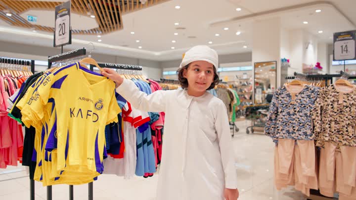 العلاقة الأسرية بين الأم والطفل أثناء التسوق، ولد عربي خليجي اماراتي يرتدي الزي التقليدي يقوم باختيار قميص أطفال ملون داخل متجر ملابس برفقة والدته، مفهوم التسوق العائلي داخل المولات التجارية، اختيار ملابس الأطفال بعناية واهتمام