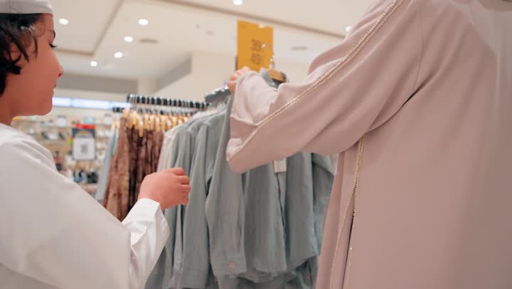 الاهتمام بالمظهر والراحة للأطفال، ولد عربي خليجي اماراتي يرتدي الزي التقليدي يقوم بتجربة ملابس أطفال داخل متجر الملابس برفقة والدته، اختيار قطعة ملابس مناسبة، مفهوم التسوق العائلي داخل المول، العلاقة الأسرية أثناء التسوق