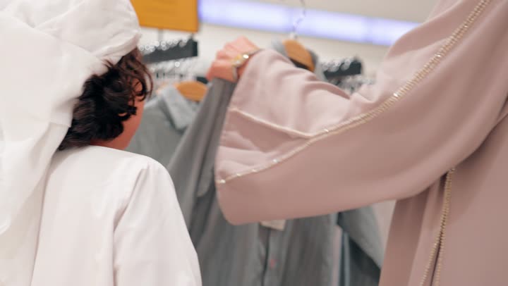 ولد عربي خليجي اماراتي يرتدي الزي التقليدي يقوم بتجربة ملابس أطفال داخل متجر الملابس برفقة والدته، اختيار قطعة ملابس مناسبة، مفهوم التسوق العائلي داخل المول، الاهتمام بالمظهر والراحة للأطفال، العلاقة الأسرية أثناء التسوق