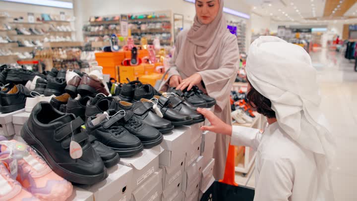 The concept of family shopping inside the mall, choosing the right shoe size, paying attention to children's comfort and the quality of shoes, an Emirati Arab boy wearing traditional attire trying on children's shoes inside a shoe store accompanied by his mother.
