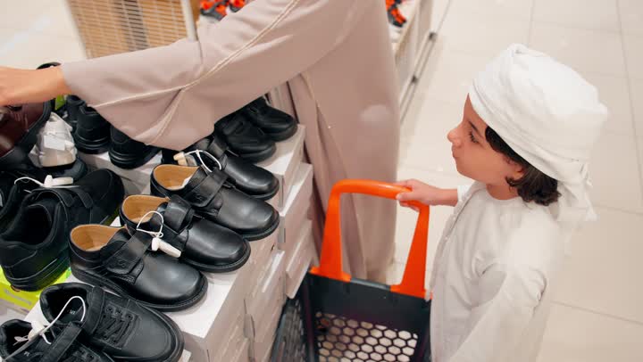 The focus on children's comfort and the quality of shoes, an Emirati Gulf Arab boy wearing traditional attire trying on children's shoes inside a shoe store accompanied by his mother, choosing the right shoe size, the concept of family shopping in the mall.