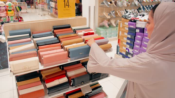 تصوير مقرب لامرأة عربية خليجيه اماراتية محجبة تقوم باختيار محفظة نسائية داخل متجر إكسسوارات ، مفهوم اختيار الإكسسوارات النسائية بعناية، التسوق داخل المولات التجارية، الأناقة النسائية والاهتمام بالتفاصيل