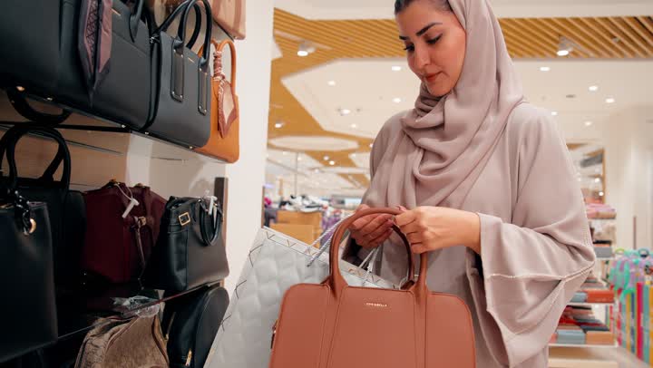 العناية بالمظهر مع إبراز الأناقة النسائية، تصوير لامرأة عربية خليجيه اماراتية ترتدي العباءة و الحجاب تقوم بتجربة حقيبة يد نسائية داخل متجر إكسسوارات، التسوق داخل المولات، مفهوم اختيار الإكسسوارات النسائية بعناية