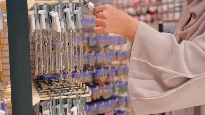 التسوق واختيار الإكسسوارات، تصوير مقرب لامرأة عربية خليجيه اماراتية ترتدي العباءة تقوم بتفقد سلاسل وعقود نسائية معروضة داخل متجر إكسسوارات، مفهوم الموضة والأناقة